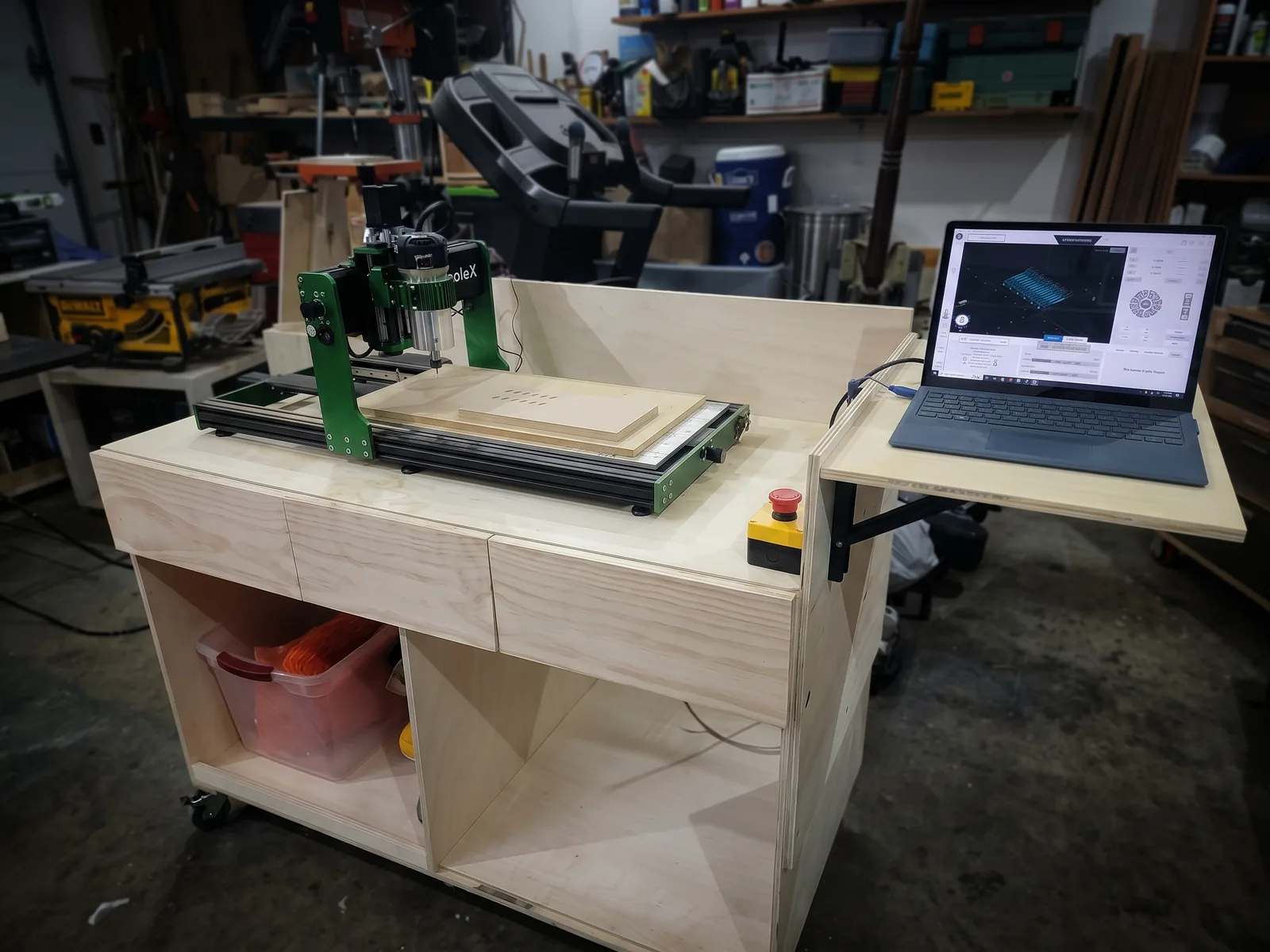 A green CNC carving machine on a plywood cart with drawers, a red stop button, and a fold-out shelf holding a laptop.